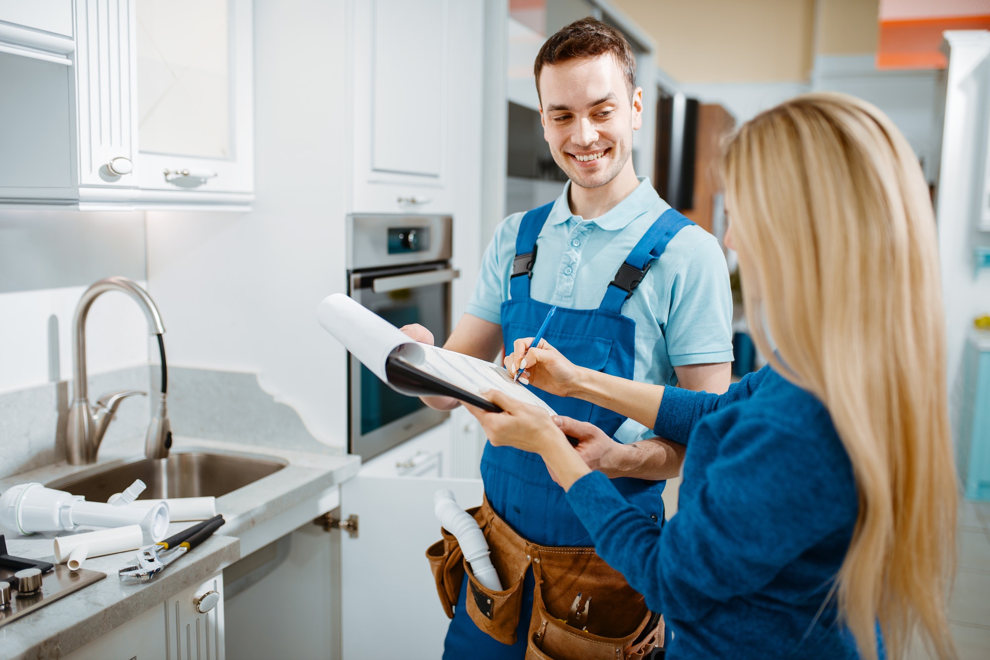 Smiling plumber in blue overalls with client in kitchen